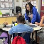 Teacher standing at a table where students are working on computers.